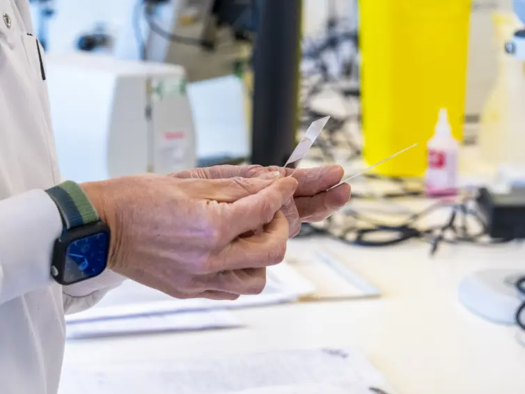 A scientist in a lab coat holds a glass slide and a thin tool in a laboratory setting.