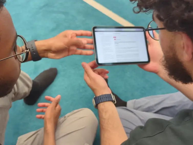 Two people sitting on a blue carpet, looking at a tablet screen and discussing something. One person holds the tablet while the other gestures.