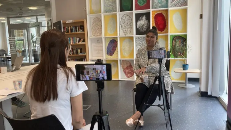 A woman in a white top is filming an interview with another woman seated in a modern, well-lit room. The camera is mounted on a tripod, capturing the seated woman The background features a colorful wall display of abstract artwork, a bookshelf, and large windows allowing natural light into the space.