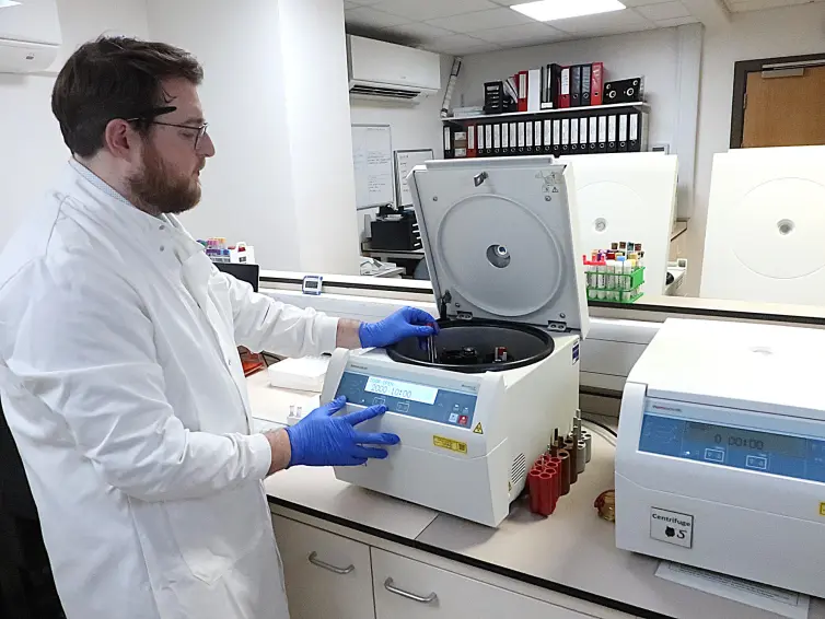 A lab manager (Andrew) in a white coat and blue gloves operates a centrifuge in a laboratory.