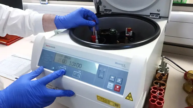 A laboratory technician wearing blue gloves and a white lab coat is handling a blood sample tube while operating a centrifuge.