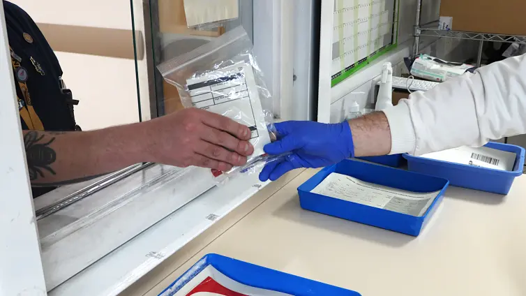 Two hands exchanging a plastic bag containing samples through a window at a laboratory facility. The hand of the right is wearing blue plastic gloves and you can see some of the lab coat.