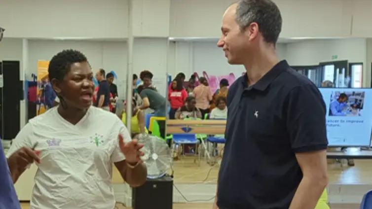 Two people, one woman speaking to a man (Bruno) in a room with other people.