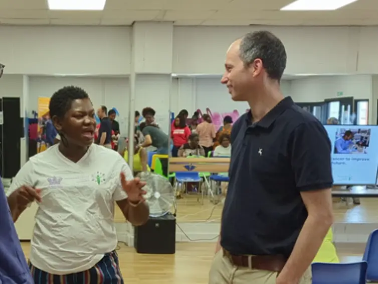 Two people, one woman speaking to a man (Bruno) in a room with other people.