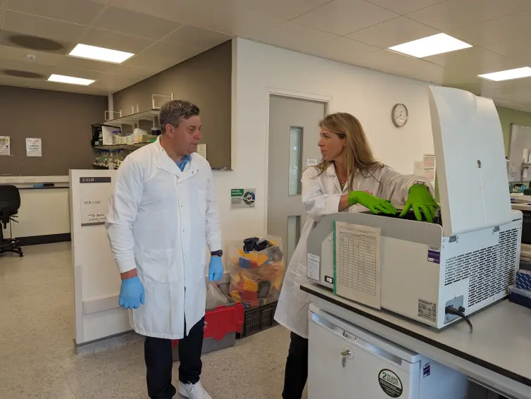 Two people wearing white lab coats in a laboratory. The male on the left, wearing blue gloves, is observing, while the female on the right, who is wearing green gloves stand by a large centrifuge machine with an open lid.