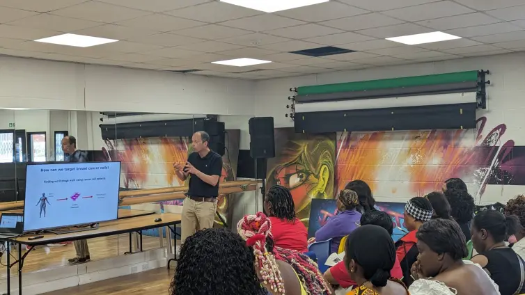 A man in a black top and beige pants stand in front of a large TV screen and presents to a room of people.