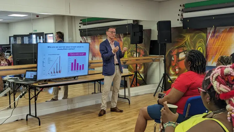 A man in a pink shirt, blue blazer and beige pants stand in front of a large TV screen and presents to a room of people.