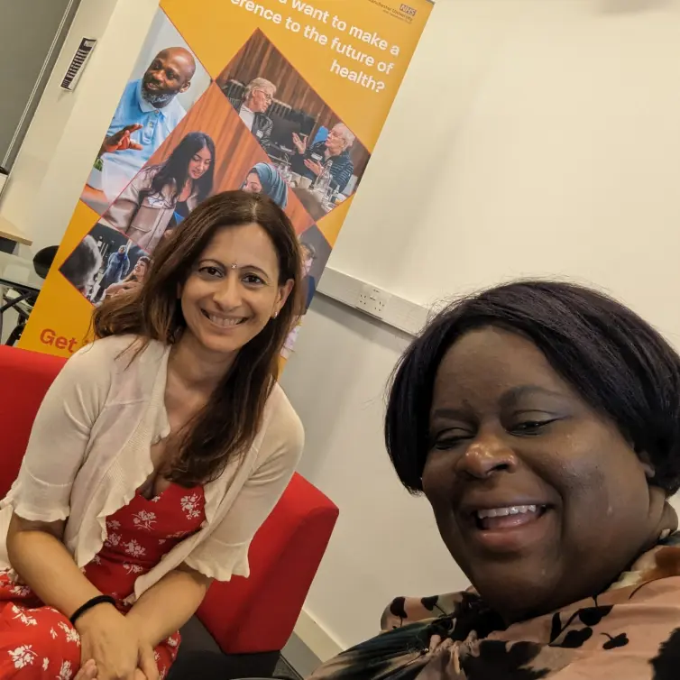 Two people posing for a selfie. On the right we have Khumbo and on the left is Maya. They're posing in front of a pull up banner saying 'Do you want to make a difference to the future of health? Get Vocal and have your say in research'.