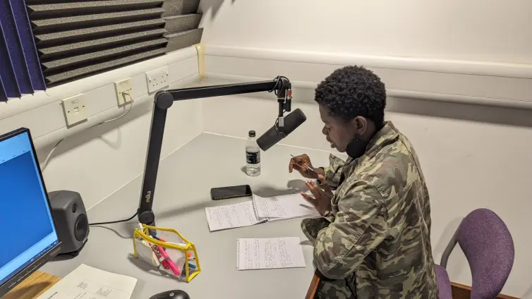 A person (Temitope) at a desk in a recording studio with a microphone, and pen and paper, looking at notes.