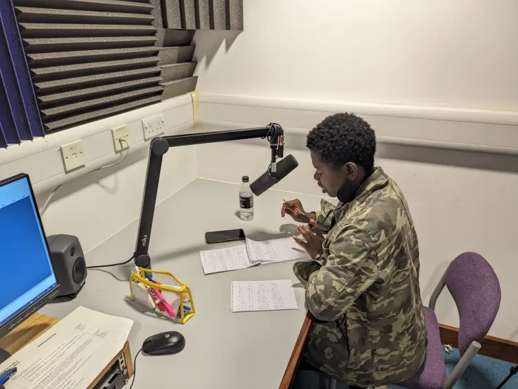 A person (Temitope) at a desk in a recording studio with a microphone, and pen and paper, looking at notes.