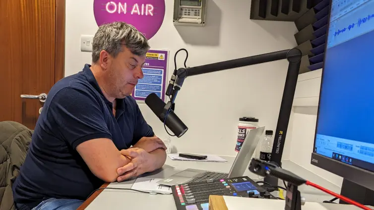 A person (Simon) at a desk in a recording studio, with a microphone, looking at a laptop screen.