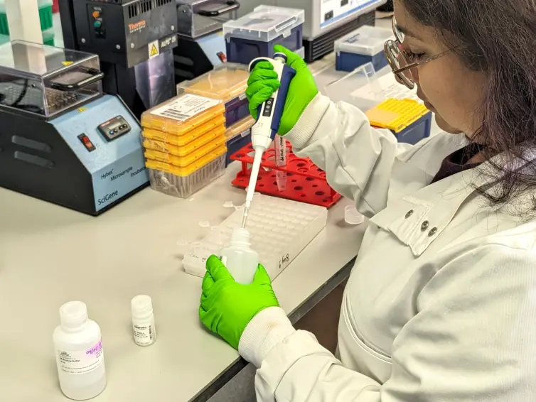 A scientist in a lab coat and green gloves is using a pipette to transfer liquid into a bottle at a laboratory workstation.