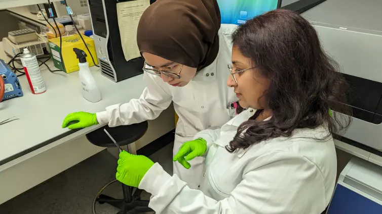 Two people wearing green gloves and white lab coats. One of them is wearing a is wearing a headscarf over covering her head. They're intent in looking at results from a test.