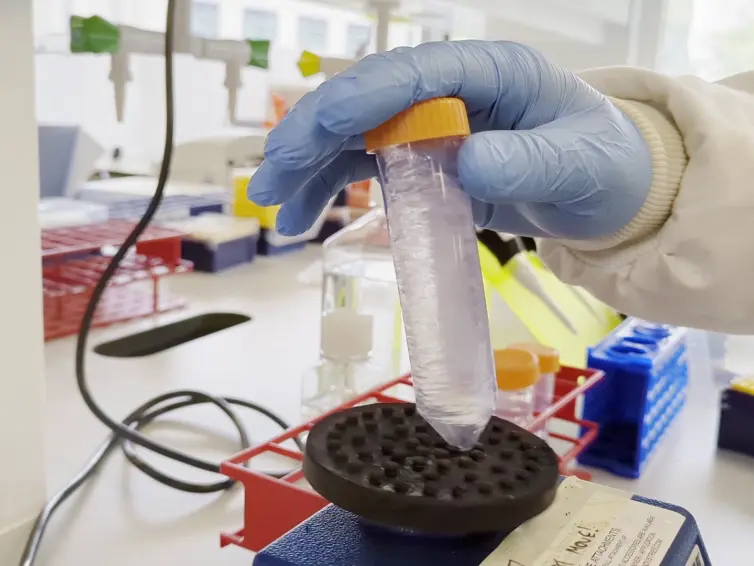 Gloved hand holds a test tube with a purple liquid, placing it on a laboratory vortex mixer, surrounded by lab equipment.