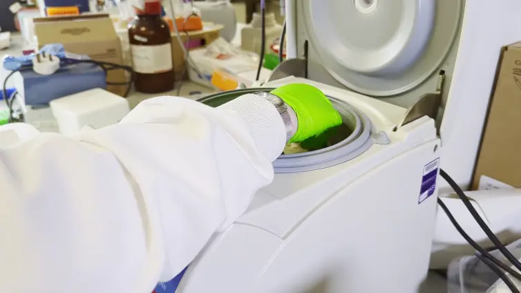 A scientist in a lab coat and green gloves is using a centrifuge machine at a laboratory workstation.