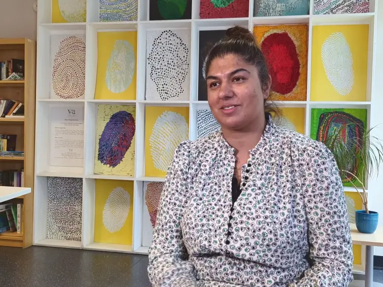 A smiling woman (Fozia) sits in front of a colorful fingerprint art display, with a bookshelf and table in the background.