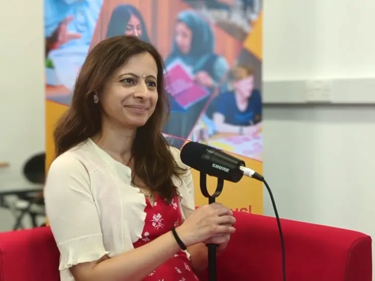 A smiling woman (Maya)  in a red floral dress sat holds a microphone while seated on a red chair.