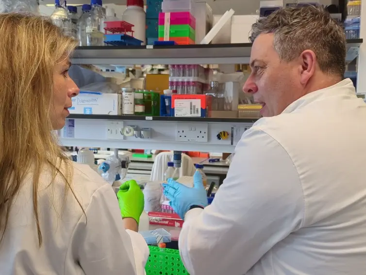 wo people in white lab coats are talking while working in a lab. One is wearing green gloves, the other blue gloves, and they are handling lab equipment. Shelves with supplies are in the background.