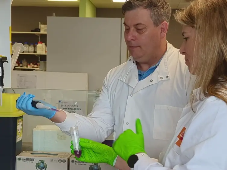 Two people wearing white lab coats. One is holding a pipette and another one is holding a tube.