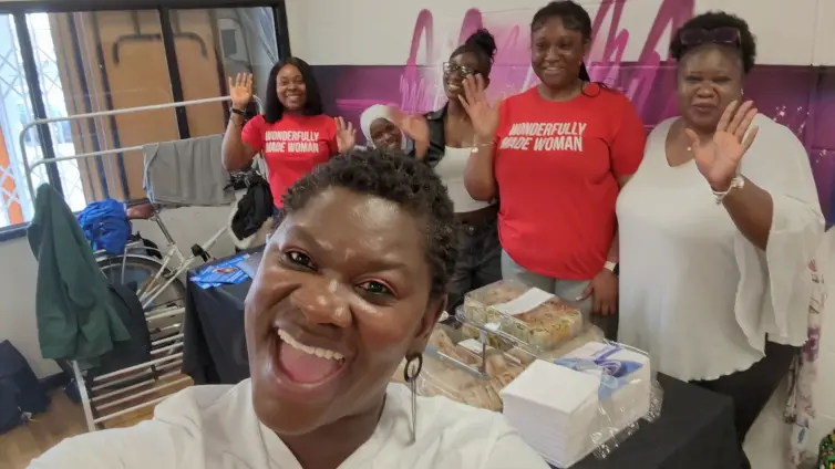 Group of women smiling and waving behind a table with food trays and supplies, one woman in front taking a selfie.