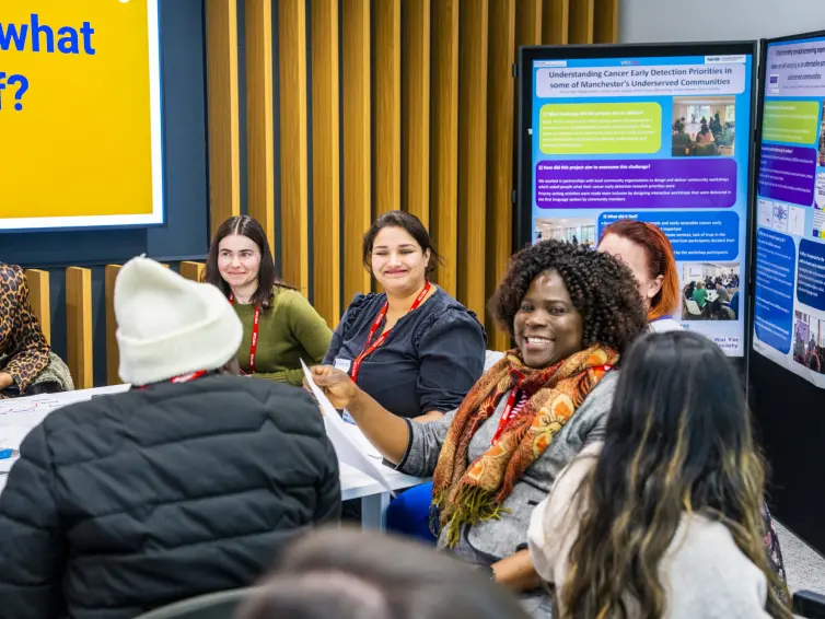 A group of diverse individuals engaged in a discussion at a round table, with informational posters in the background. Everyone is smiling, a Black woman is looking at the camera.