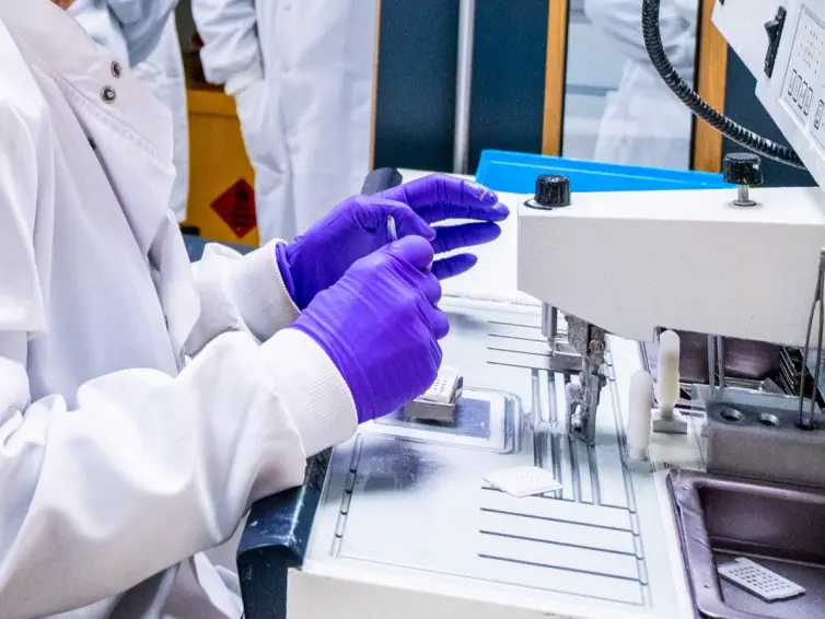 A researcher wearing a white lab coat and purple gloves handles a small sample next to a piece of laboratory equipment. A second person in a white lab coat is visible in the background.