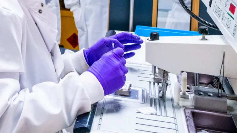 A researcher wearing a white lab coat and purple gloves handles a small sample next to a piece of laboratory equipment. A second person in a white lab coat is visible in the background.