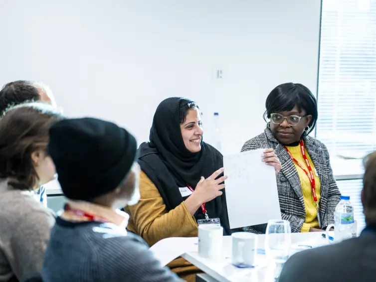 Photograph of a diverse group of people engaged in a discussion around a table in a bright room. One woman wearing a black headscarf and mustard sweater holds up a sheet of paper while others listen attentively, with cups, glasses, and water bottles visible on the table.