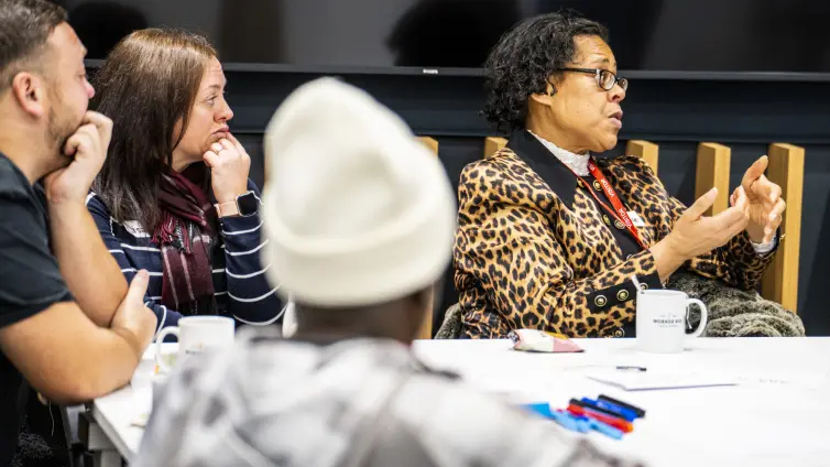 Photograph of a group discussion with four people seated around a table, focusing on a woman in a leopard print jacket speaking and gesturing with her hands. Table holds coffee mugs, papers, and colorful markers, indicating a collaborative or brainstorming session.