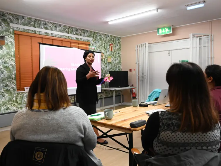 A person in a black outfit is presenting to a group in a room with leafy wallpaper and wooden blinds.
