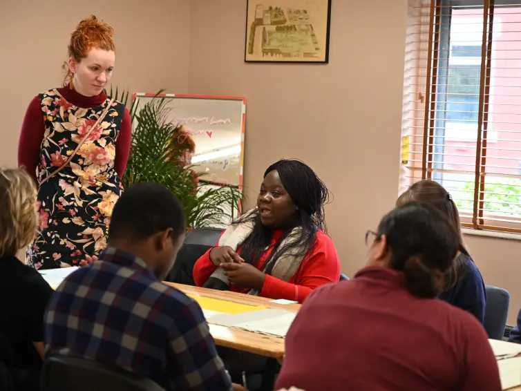 A group of people sit around a table during a meeting. One woman in a floral dress stands, while another in a red sweater speaks.