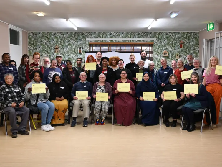 A diverse group of people stands and sits in a community room, smiling and holding certificates. The room has light walls and a leafy wallpaper.