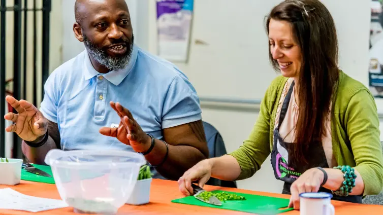 Photograph of two people engaged in a cooking activity at a table with cutting boards, chopped vegetables, and a clear mixing bowl. Man in a light blue shirt gestures while talking, and woman in a green cardigan smiles while preparing food, suggesting a collaborative and enjoyable cooking session.