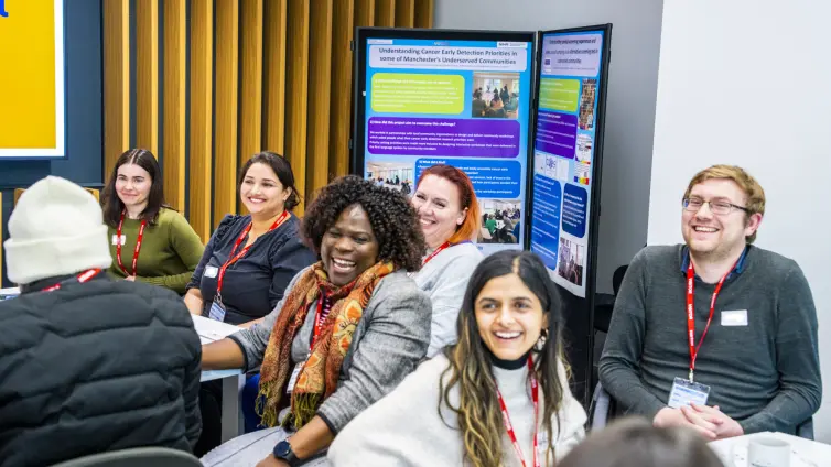 A group of people seated in a room, engaged in a discussion or meeting. Posters or presentation boards with text and images are visible in the background.