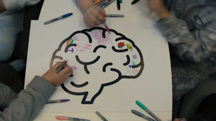 Three people are colouring and doodling on a large outline of a brain, using colourful markers. Various symbols and small drawings decorate the brain, and additional markers are scattered around the paper.