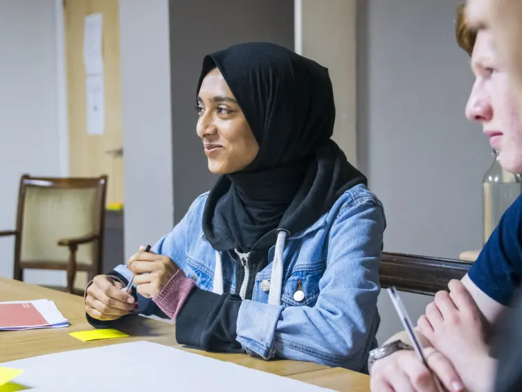 A woman in a hijab sits at a table with two other people, engaged in conversation.