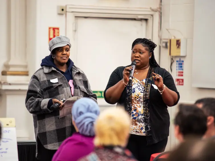 Two women stand at the front of a room; one holds a clipboard while the other speaks into a microphone to an audience.