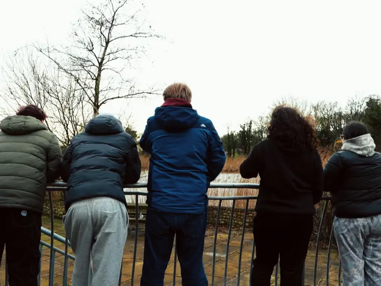 Five young people wearing winter jackets stand with their backs to the camera, leaning on a metal railing and looking out over a grassy landscape with bare trees on a cloudy day.
