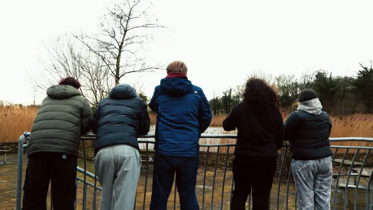 Five young people wearing winter jackets stand with their backs to the camera, leaning on a metal railing and looking out over a grassy landscape with bare trees on a cloudy day.