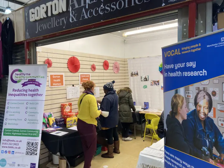 People visit informational stalls with banners about health research and community health at an indoor event, with red decorations on the wall.