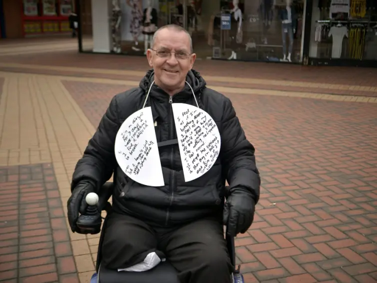 Man in wheelchair in street with board around his neck with his experiences of breathlessness written on it.