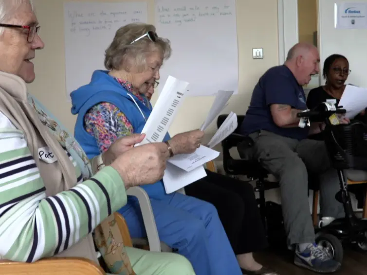 Image of four people sitting down singing with song sheets in hands.