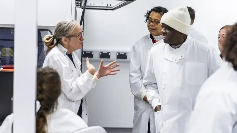 A group of people from Manchester wearing white lab coats engage in conversation with a scientist who is gesturing with her hands while others listen attentively.