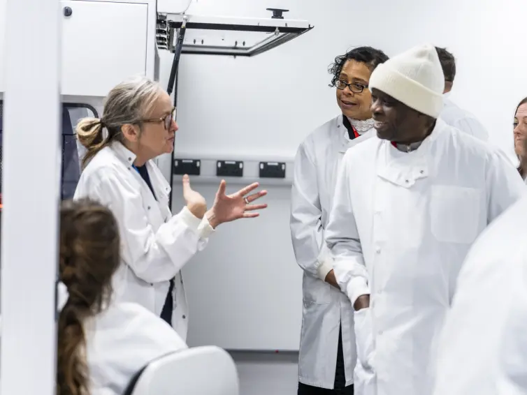 A group of people from Manchester wearing white lab coats engage in conversation with a scientist who is gesturing with her hands while others listen attentively.