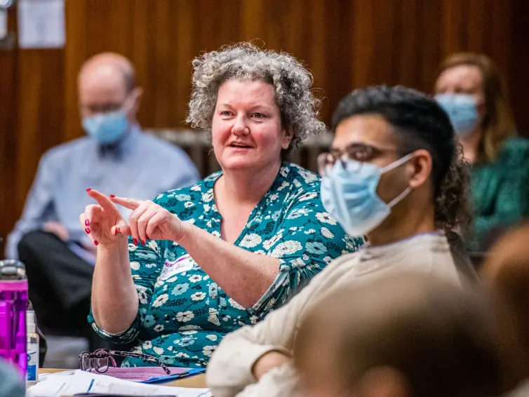 A female presenting person talking to someone off camera, someone is in the foreground listening whilst wearing a surgical mask.