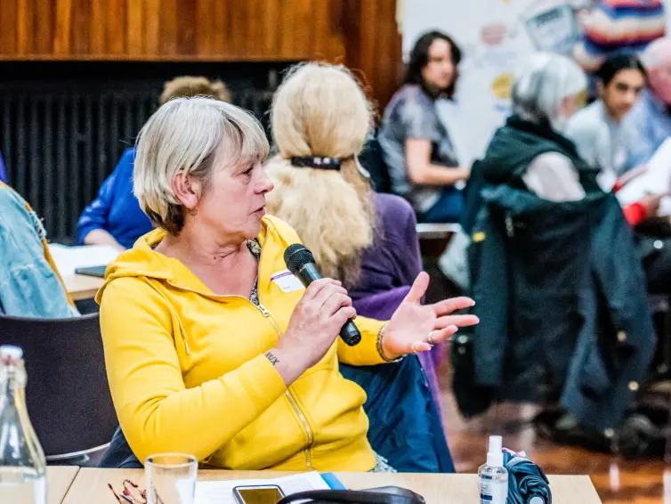 Person in yellow top talking into a microphone and gesturing in a busy room.