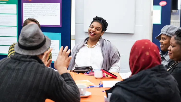 People sitting around a table, engaged in a friendly discussion. A woman in the center is smiling, and others are actively participating.