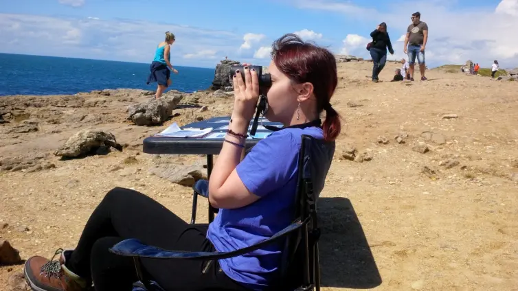 Young person sitting on a chair on a cliff, looking at the sea with binoculars.