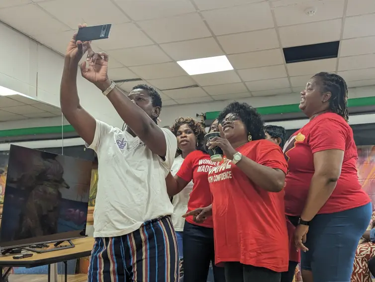 A group of five women stands close together at a woman to discuss laboratory research and how it benefits Black African women with breast cancer and prevention; one woman on the left is taking a selfie while the others smile. Two women are wearing red shirts,, while the rest are in casual clothing.