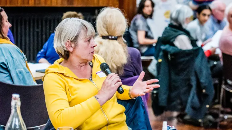 A person wearing a yellow hoodie sat down at a table and talking with a microphone.